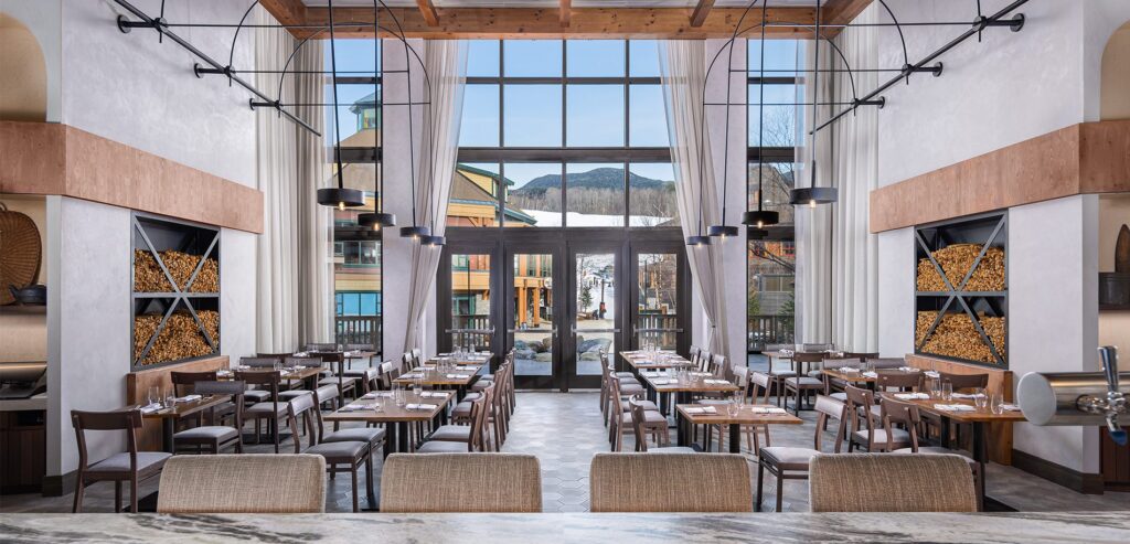 The interior of an empty restaurant with high ceilings and large windows looking out into a mountain resort.