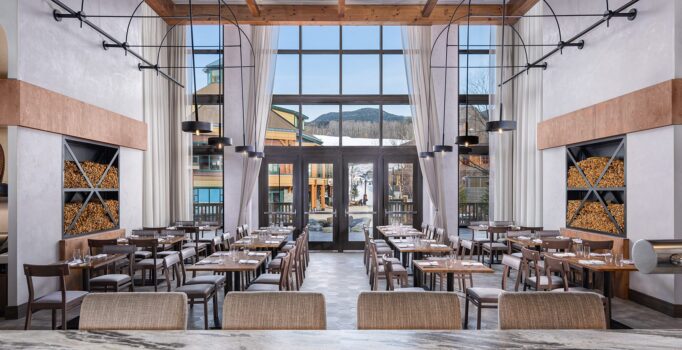The interior of an empty restaurant with high ceilings and large windows looking out into a mountain resort.