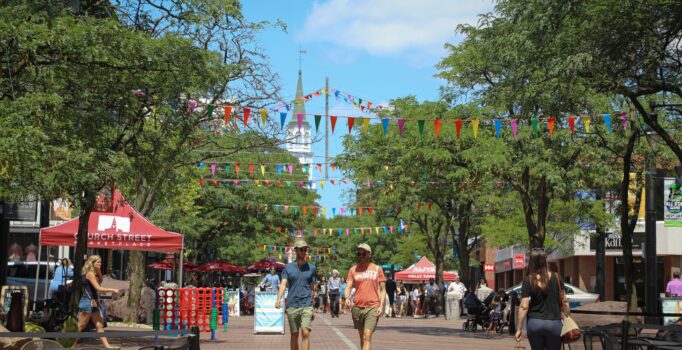Pedestrians walk down a brick marketplace on a sunny day.