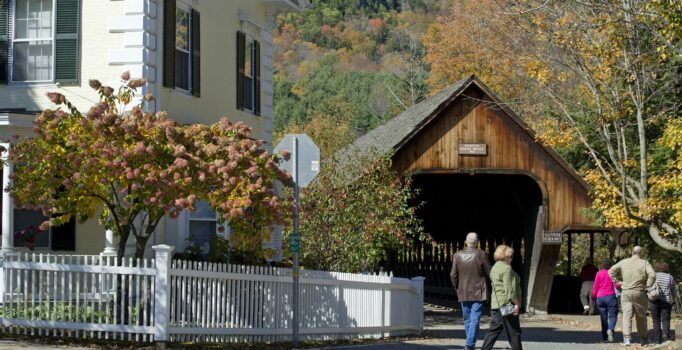 A group of people walk towards a brown covered bridge in downtown Woodstock.