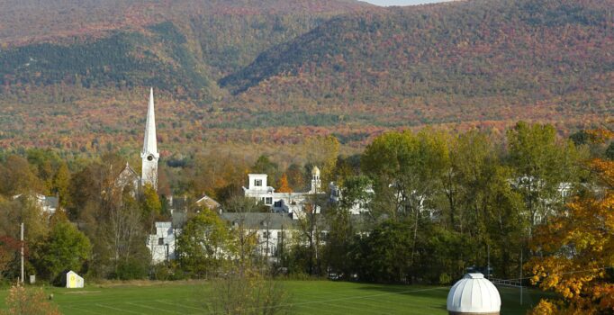 A town skyline is visible above trees with fall foliage surrounded by mountains.