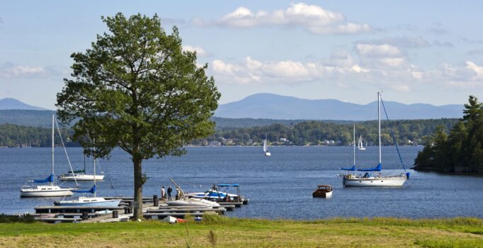 A large lake, surrounded by mountain peaks and full of boats, is seen on a sunny day.