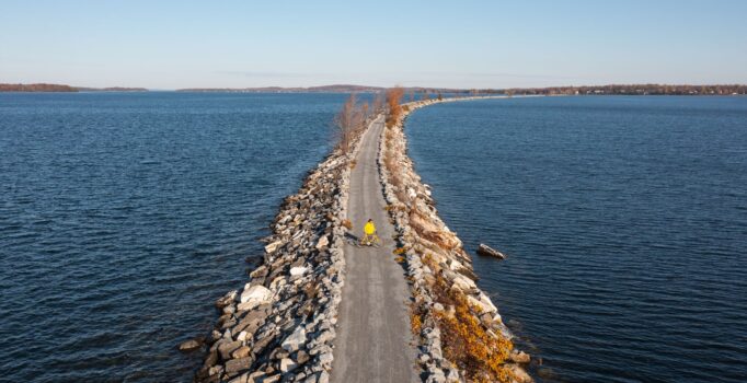 A person seen from far above with a bike on a gravel trail crossing a blue lake on a sunny day.