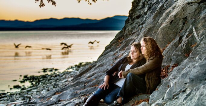 Two people lay on the rocky shoreline of a lake at dusk.