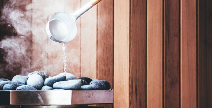 Steam billows as water is poured from a spoon onto hot rocks in a sauna.