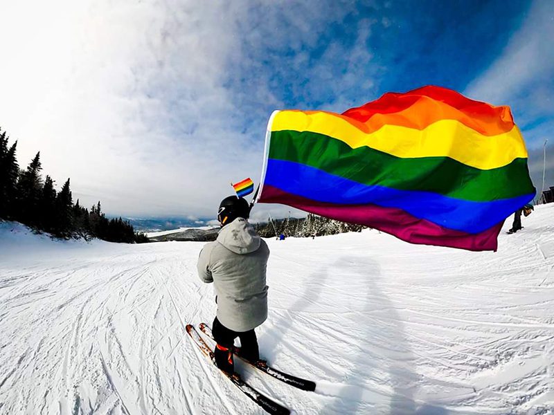 A person skis down a mountain holding a rainbow flag in the sun.