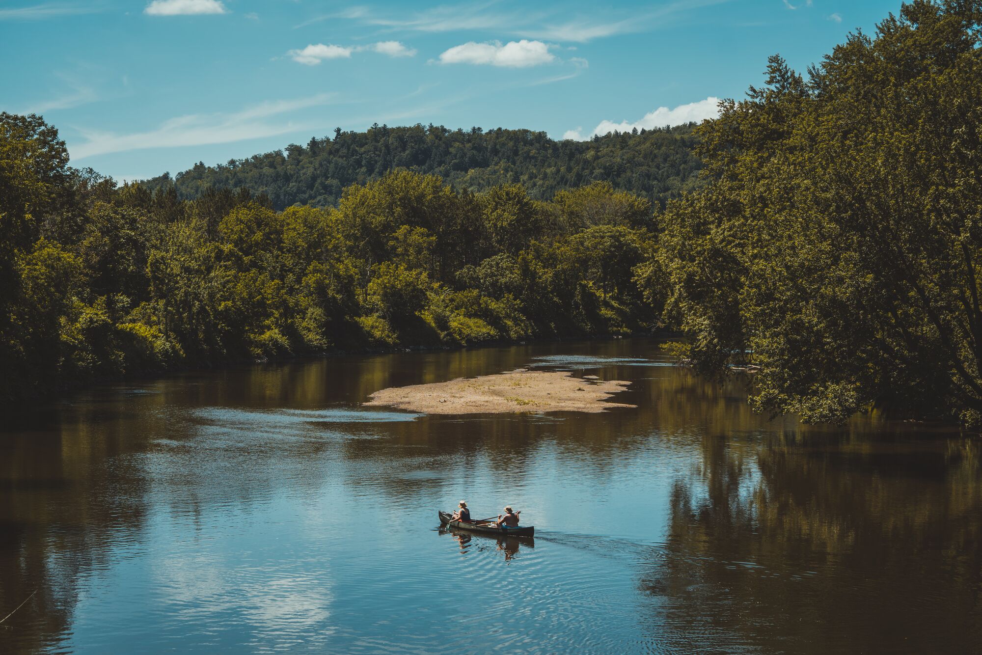 Two people paddling a canoe down a wide, calm river framed by dense green trees and a wooded hill under a bright sky.