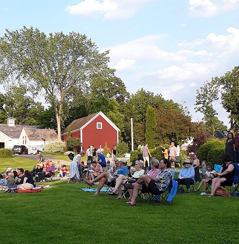 A crowd of people sit outside on a green lawn on a warm sunny day.