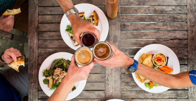 View from above looking down at three people toasting beverages over a table with four plates of food.