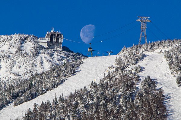 A snowy mountain is seen from below with ski trails and chairlifts to the top. The moon is visible in a blue sky.