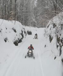 Two people snowmobile down a snowy path.