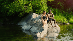 Two people sit on a large rock next to a river.