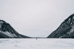 Seen from afar, a small ice fishing shanty on a frozen lake at sunset with mountains rising up on either side.