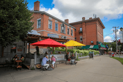 People sit at tables under canopies outside beside old brick buildings on a warm day.