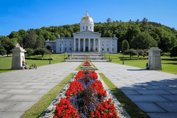 Two paved sidewalks with red flowers lead to a large white stone building with a golden Rotunda on a sunny day.