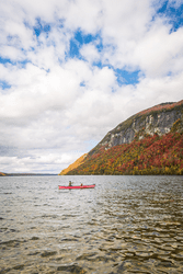 A person kayaks on a lake with colorful foliage on a mountain in the background.