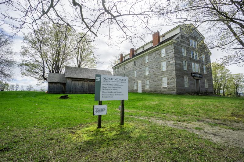 A large, multi-story historic stone building and a weathered wooden barn sit on a grassy hill behind a museum welcome sign.