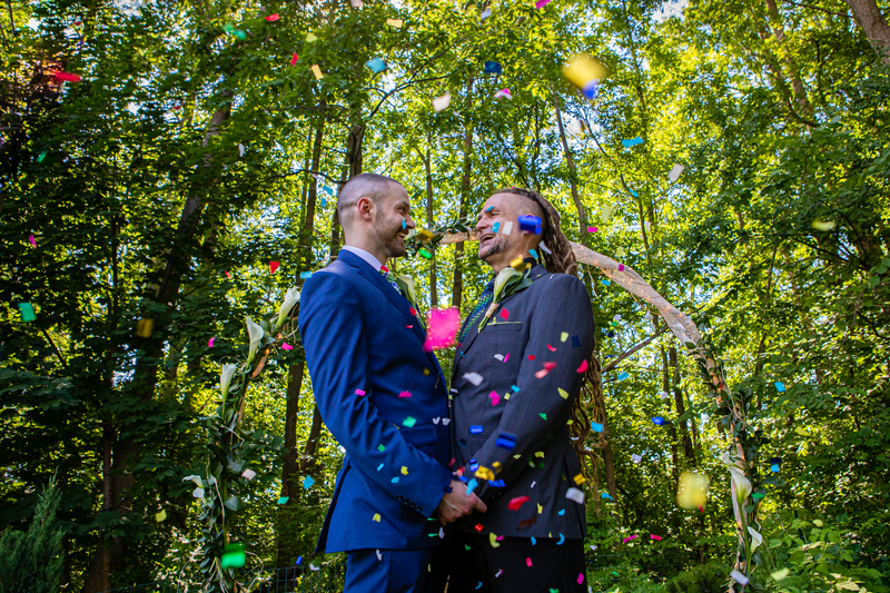 Two people in suits hold hands facing each other and smile as they get married outdoors. Confetti swirls around them.