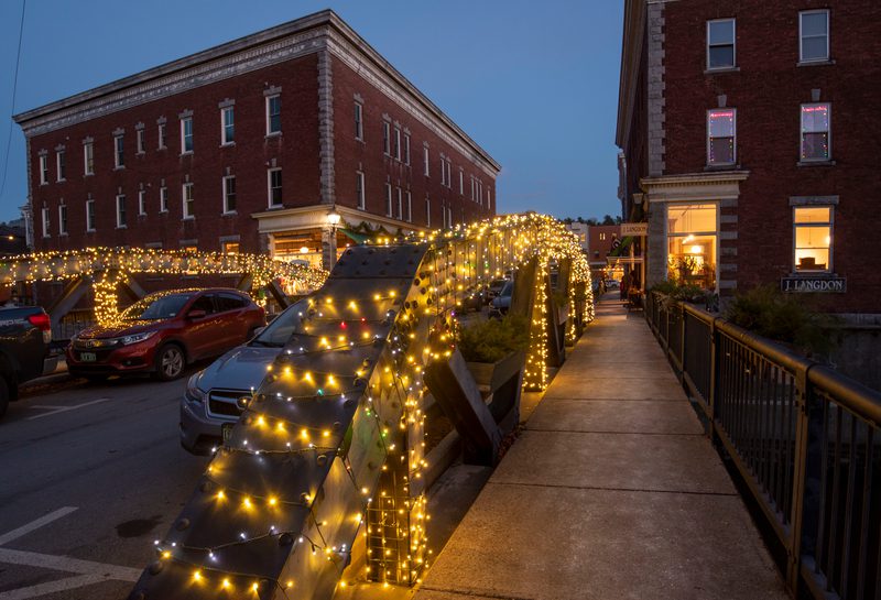 At dusk, a bridge across a river is illuminated by warm glowing lights. A historic brick building is visible in the background.