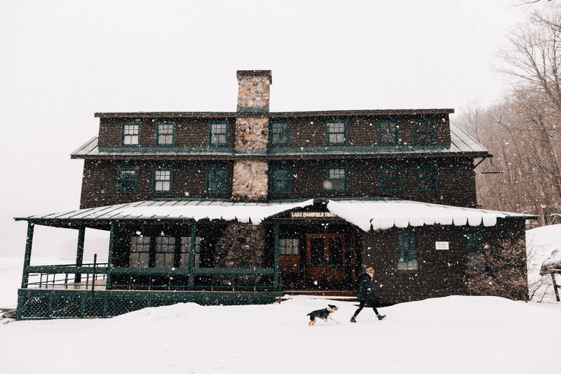 A woman walking a dog during snowfall, in front of the Lake Mansfield Trout Club.