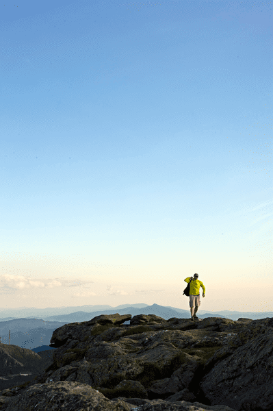 A solo hiker at the summit throwing their backpack on their shoulder, walking toward the camera, showing layers of mountain views in the background.