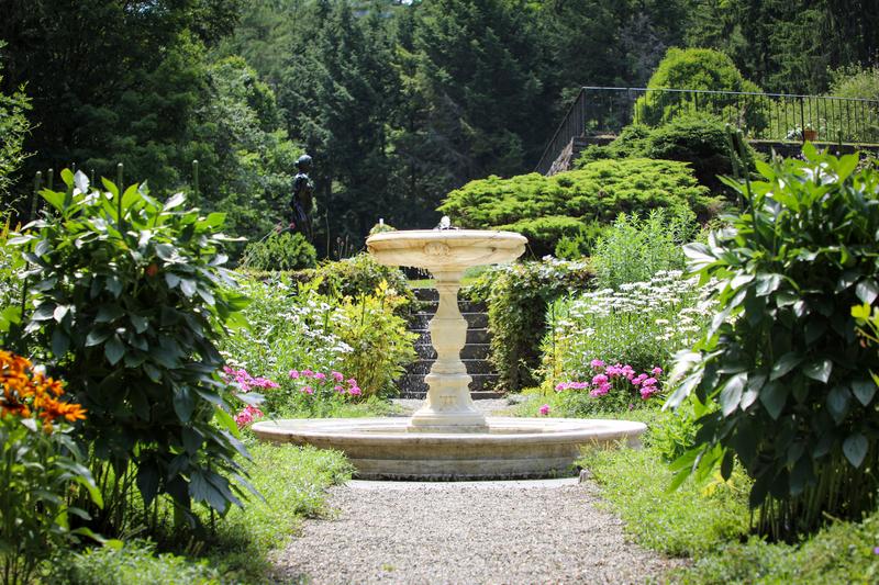A multi-tiered stone water fountain stands in the center of a lush garden surrounded by green bushes, colorful flowers, and tall trees.