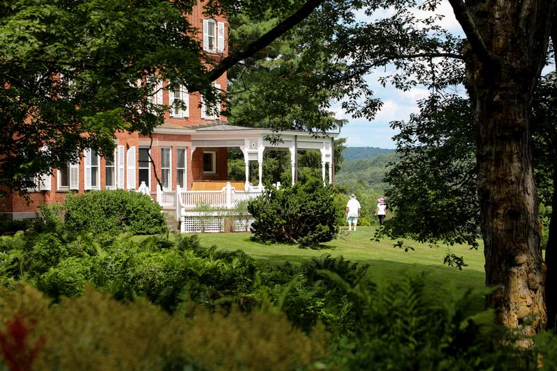 Two people walk across a sunny green lawn next to a historic brick estate with a white covered porch.