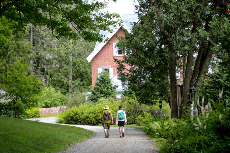 Two people wearing backpacks walk along a gravel path toward a multi-story brick house surrounded by lush trees and greenery.
