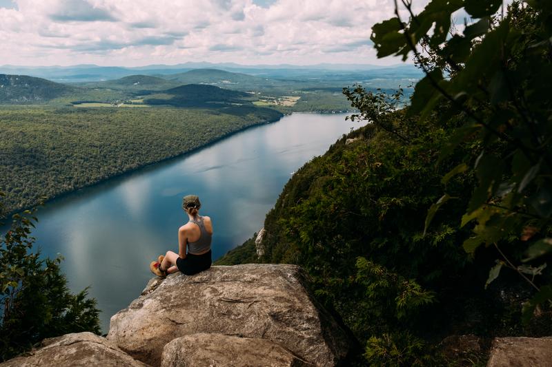 A hiker sits on a rocky cliff ledge overlooking a long, narrow lake surrounded by densely forested mountains and valleys.