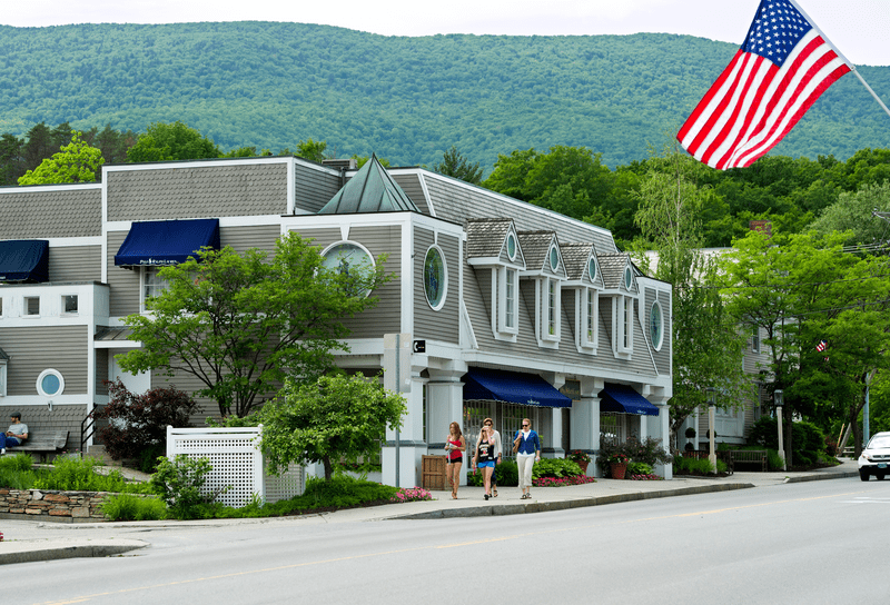 Stores line a paved road with pedestrians walking and an American flag hanging in the foreground.