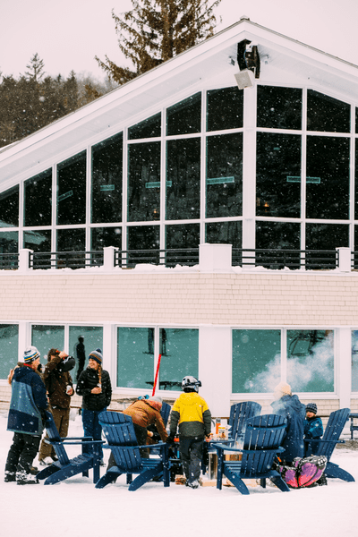 The exterior of a ski lodge welcomes visitors around a firepit during snowfall.