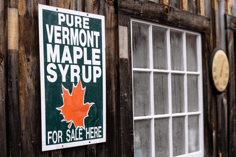 A sign reading Pure Vermont Maple Syrup For Sale Here is posted on the exterior of a wooden building.