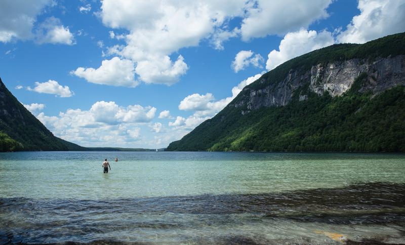 Two people wade in the clear, shallow water of a lake framed by steep, forested mountains under a partly cloudy blue sky.