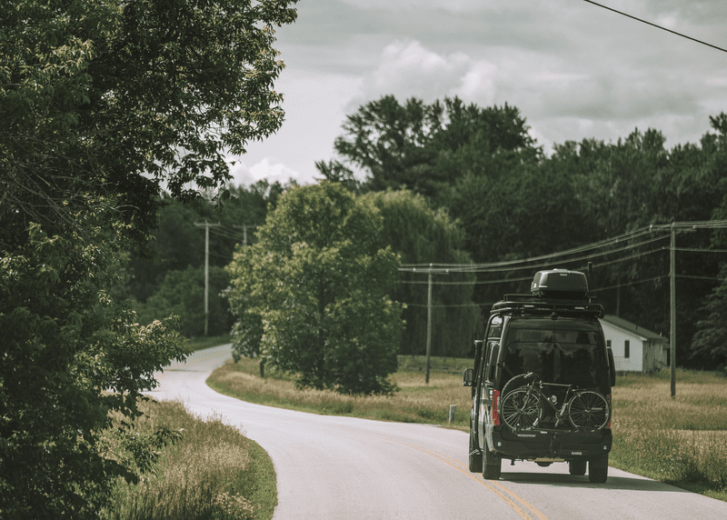 A van with bikes on the back drives on a winding road in the summer.
