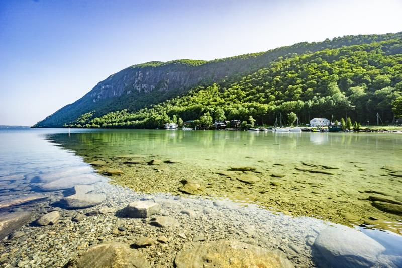 Clear, shallow water with a rocky shoreline looks out toward a steep, forested mountain rising above a lake.