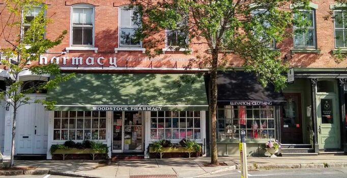 A street view of a quaint downtown area with storefronts, pedestrians, and cars parked along the road.