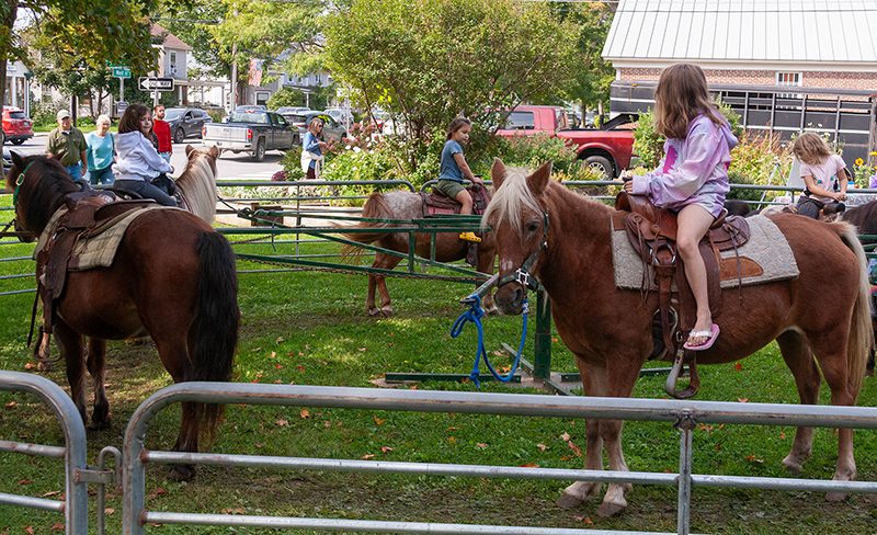 Several children ride ponies in a pen on a warm day.