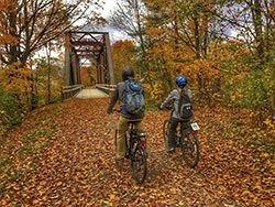 Two people seen from behind bike on a rail trail crossing a railroad bridge in the fall.