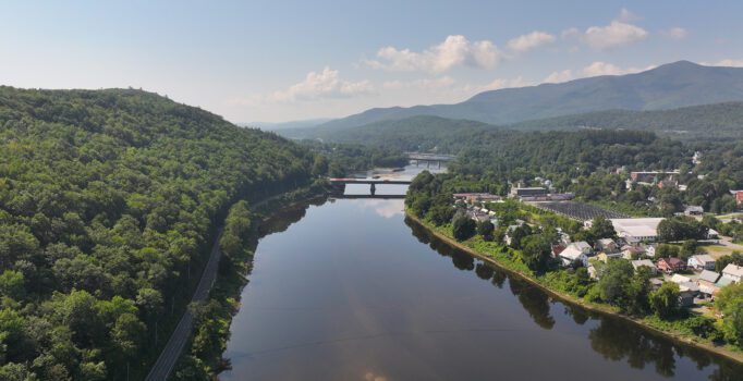 Seen from above, a covered bridge stretches across a river in the summer with a long road to the left and a town to the right.