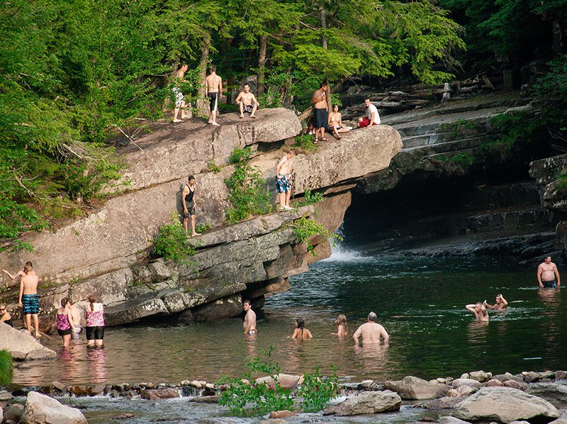 People stand on a cliff jutting out into a busy swimming hole.