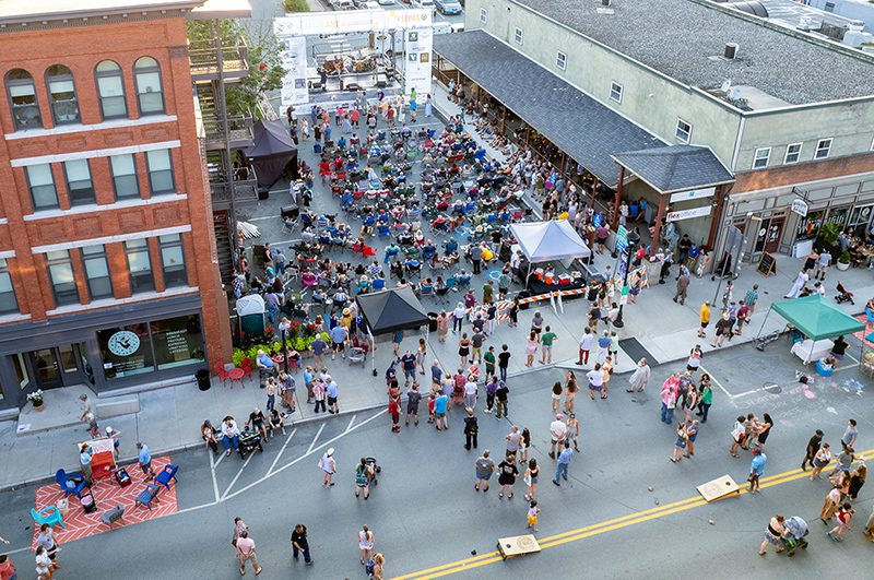 Seen from above, crowds gather on the street of a historic downtown. Tents house food vendors and live musicians.