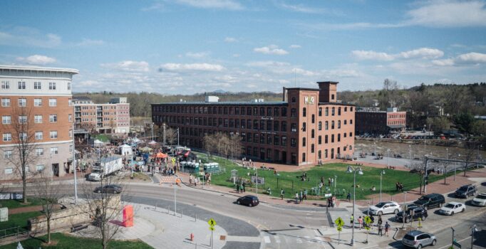 Seen from above, several large brick buildings sit beside a river and a paved road on a sunny day.