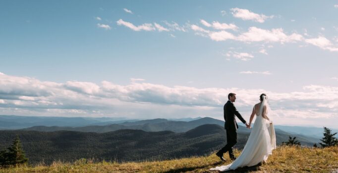 Two people in wedding attire, walk across a grassy field on a sunny day.