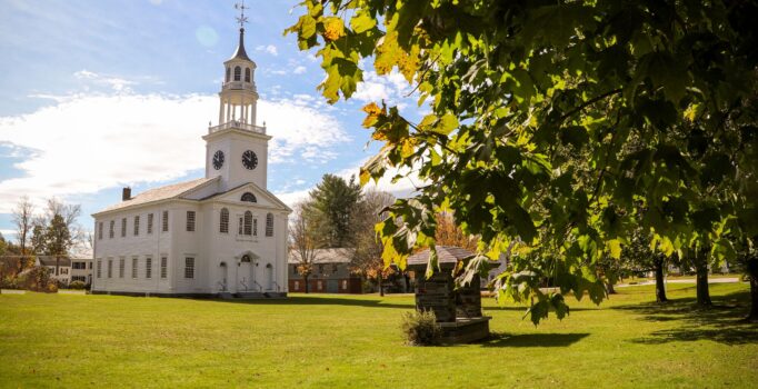 A white church on a green lawn seen from the road with trees surrounding.