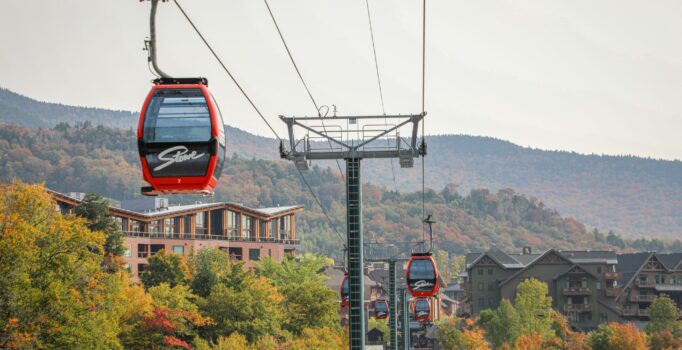 Red Stowe gondolas move across a track high above the ground. Yellow and orange trees are on the ground below.