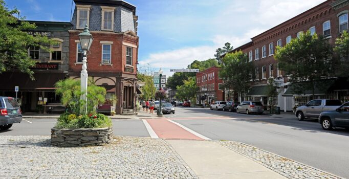 Pedestrians walk along a sidewalk among storefronts in a town center on a sunny day.