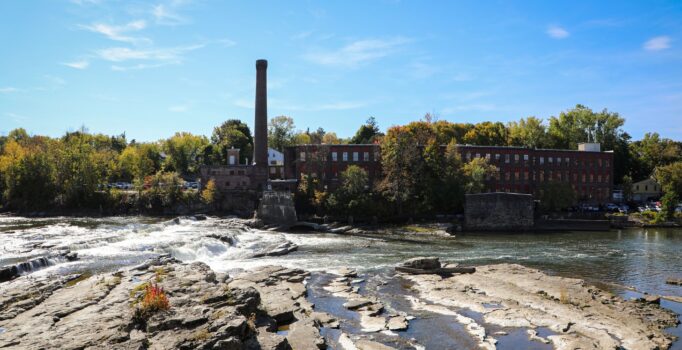 A river flows over rocks with a historic mill village in the background. 