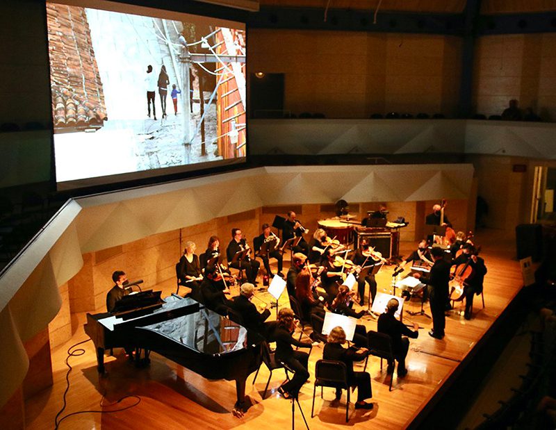 An orchestra plays on stage while a movie is projected on a screen behind them.