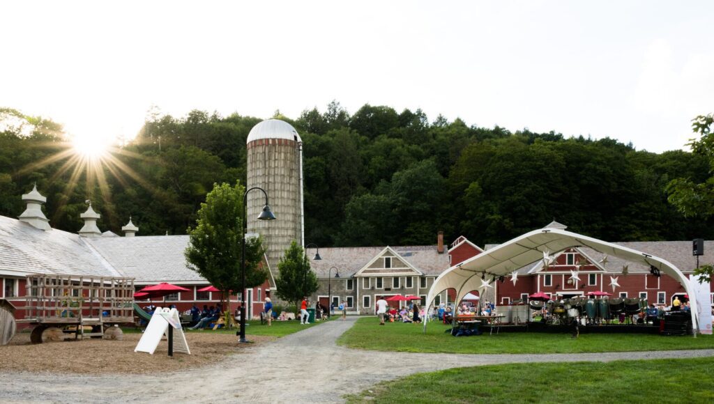 A lively outdoor gathering at a farm with a tall silo and rustic buildings. People are sitting on blankets and chairs, enjoying a sunny day amidst greenery.