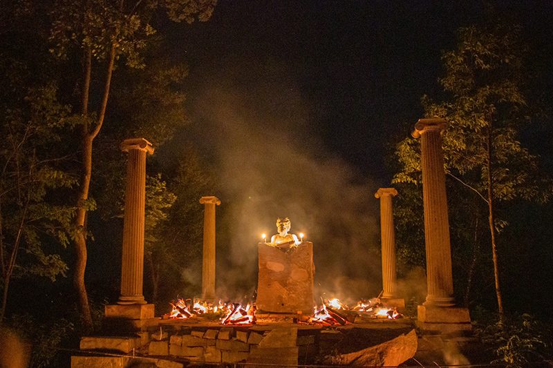 An outdoor fire burns among large pillars in the woods at night.